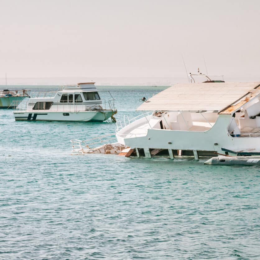 A white boat resting in a harbor, surrounded by other boats in the background. The boat appears to have suffered damage and is partially submerged in the water. It has a flat roof and a cabin area that has collapsed partially. The boat leans to the left, and its front is submerged in the light blue water. The sky above is a hazy shade of blue, adding to the overall scene.