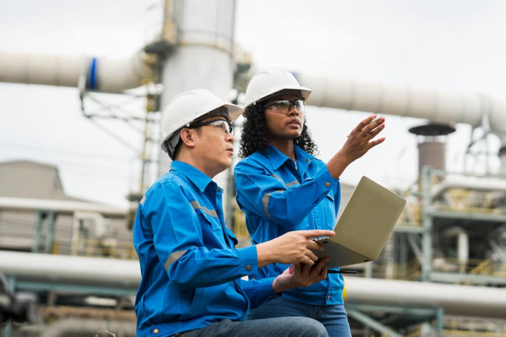 Male and female engineers work using laptop and tablet in gas refineries and oil refinery plants to check production quality and safety of refineries.