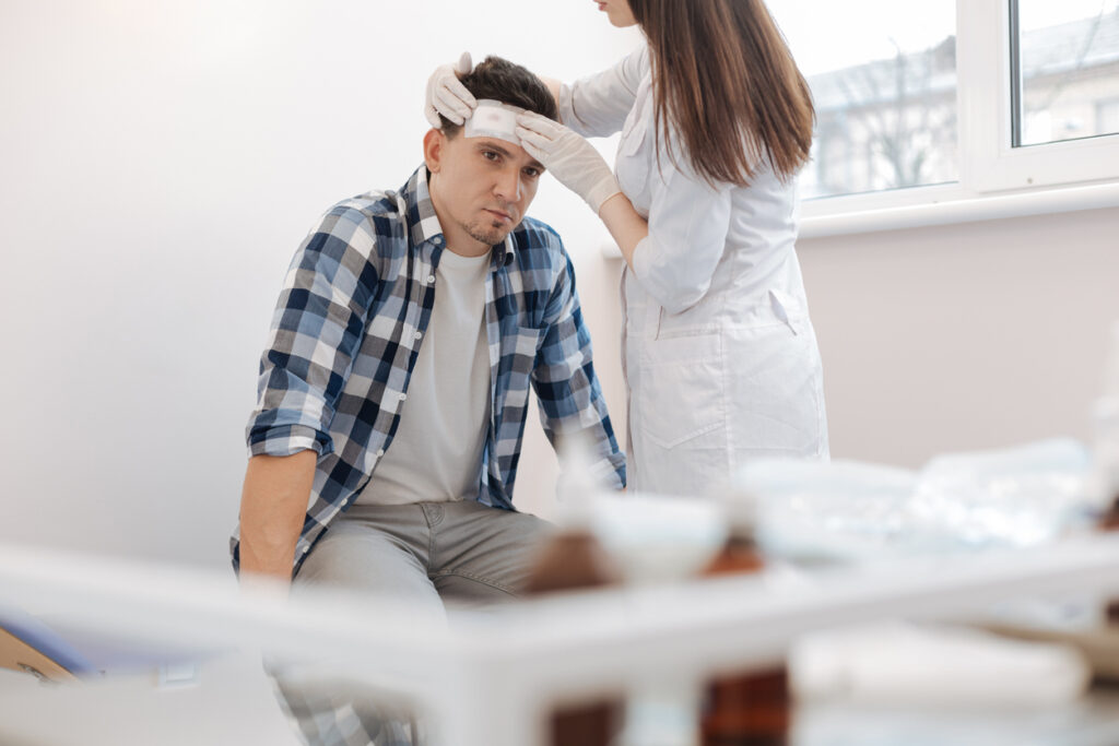 man sitting on the medical bed and receiving the first aid while having the forehead wound